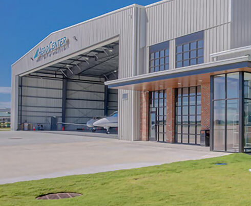 Exterior view of a large hangar with an open door, revealing small aircraft inside, set against a clear blue sky.
