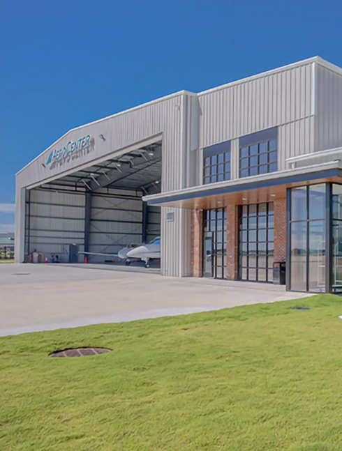 Exterior view of a large hangar with an open door, revealing small aircraft inside, set against a clear blue sky.