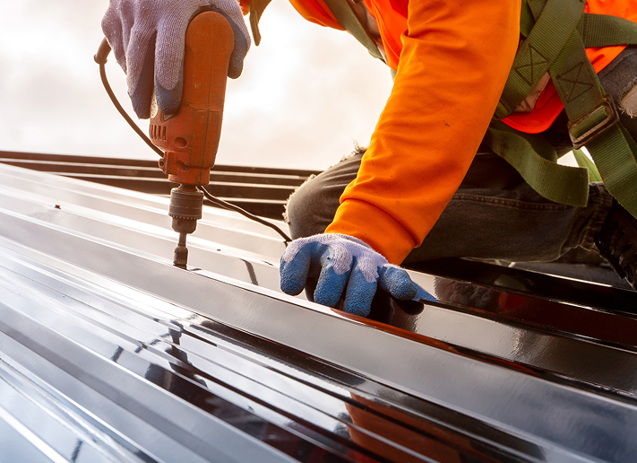 Person in safety gear using a power drill on a metal roof panel