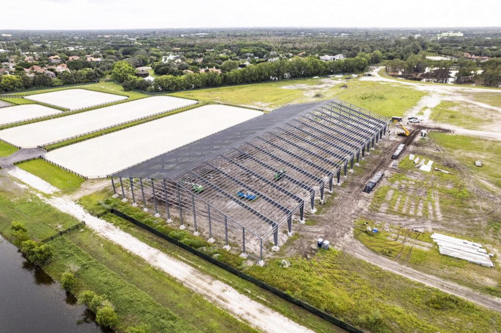 Aerial view of large construction site with steel framework near equestrian arenas and surrounding greenery.