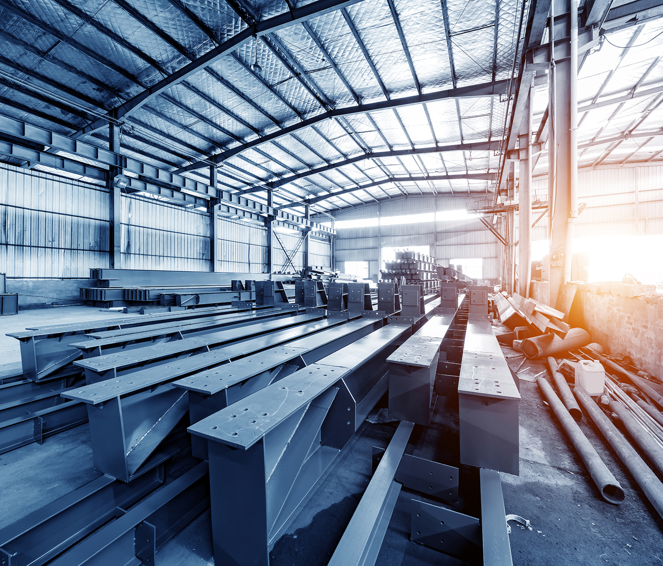 Inside a spacious factory warehouse with large steel beams organized on the floor, under a high metal ceiling.
