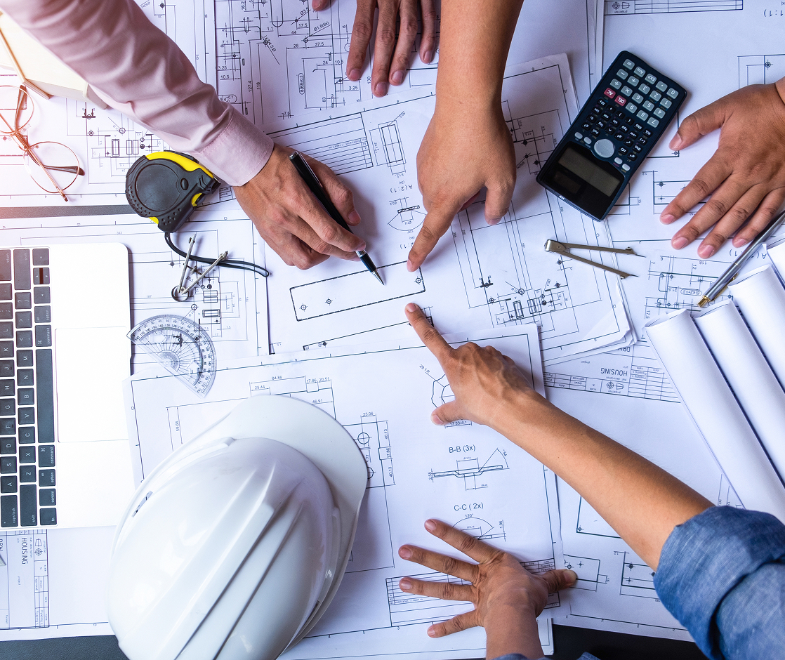 Hands collaborate over architectural blueprints with calculator, tape measure, and hard hat on desk.