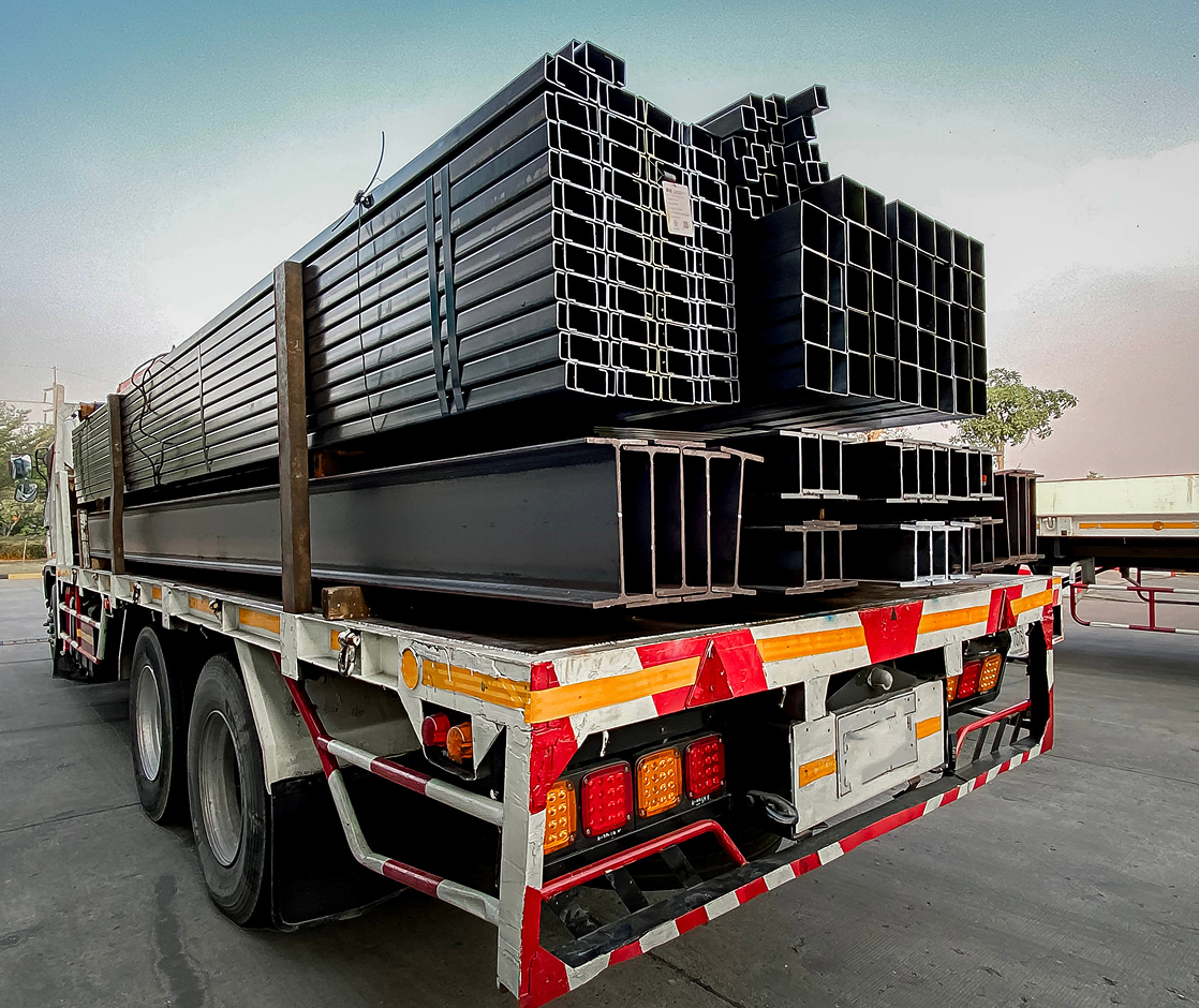 Truck loaded with various steel beams and pipes, parked on a concrete surface under a clear sky.