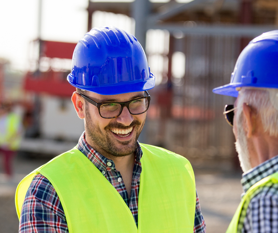 Smiling construction workers wearing blue hard hats and yellow vests talking on construction site.