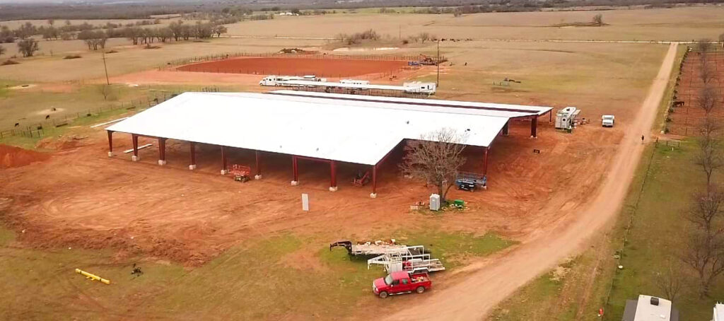 Farm building under construction on red soil, surrounded by fields and dirt road, with vehicles and equipment nearby.