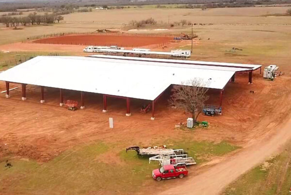 Farm building under construction on red soil, surrounded by fields and dirt road, with vehicles and equipment nearby.