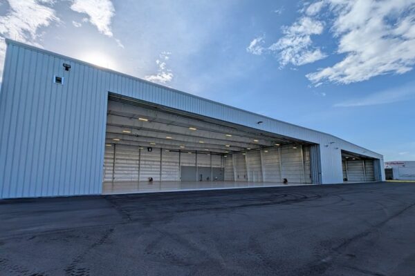Angled exterior view of a pre-engineered steel airplane hangar at Skybridge Miami Opa-Locka Airport with both hangar doors open and the lit interior visible